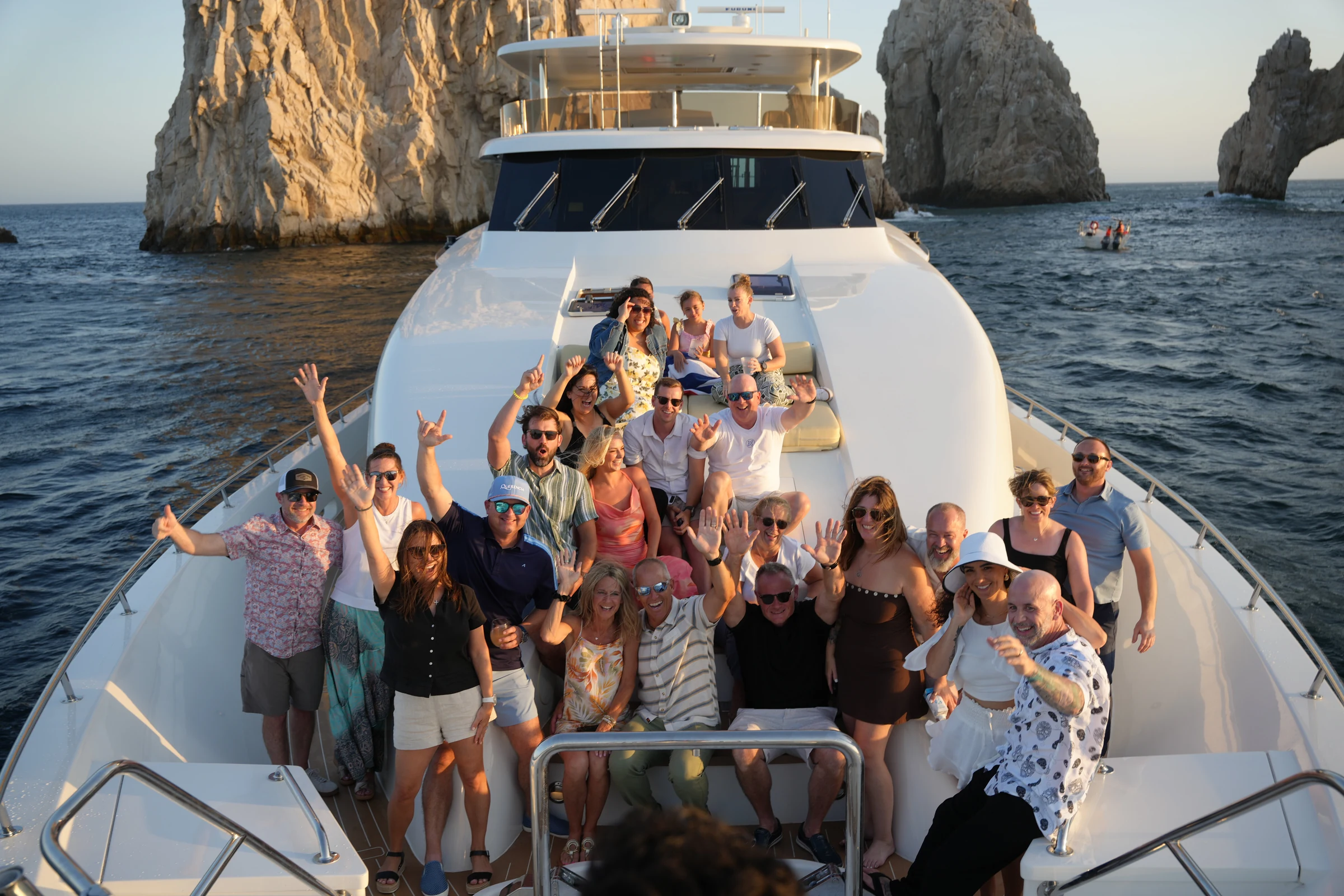 A large group smiling and waving together on a yacht near the Cabo arch